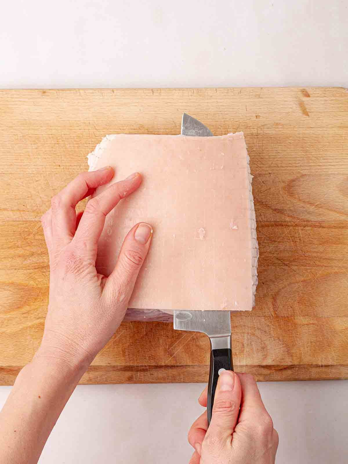 Looking down onto a pair of hands slicing off the fat of a gammon joint on a wooden chopping board.