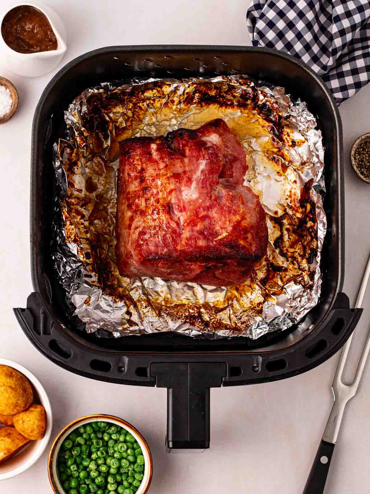 An air fryer with a cooked gammon joint inside, surrounded on table by utensils and condiments.