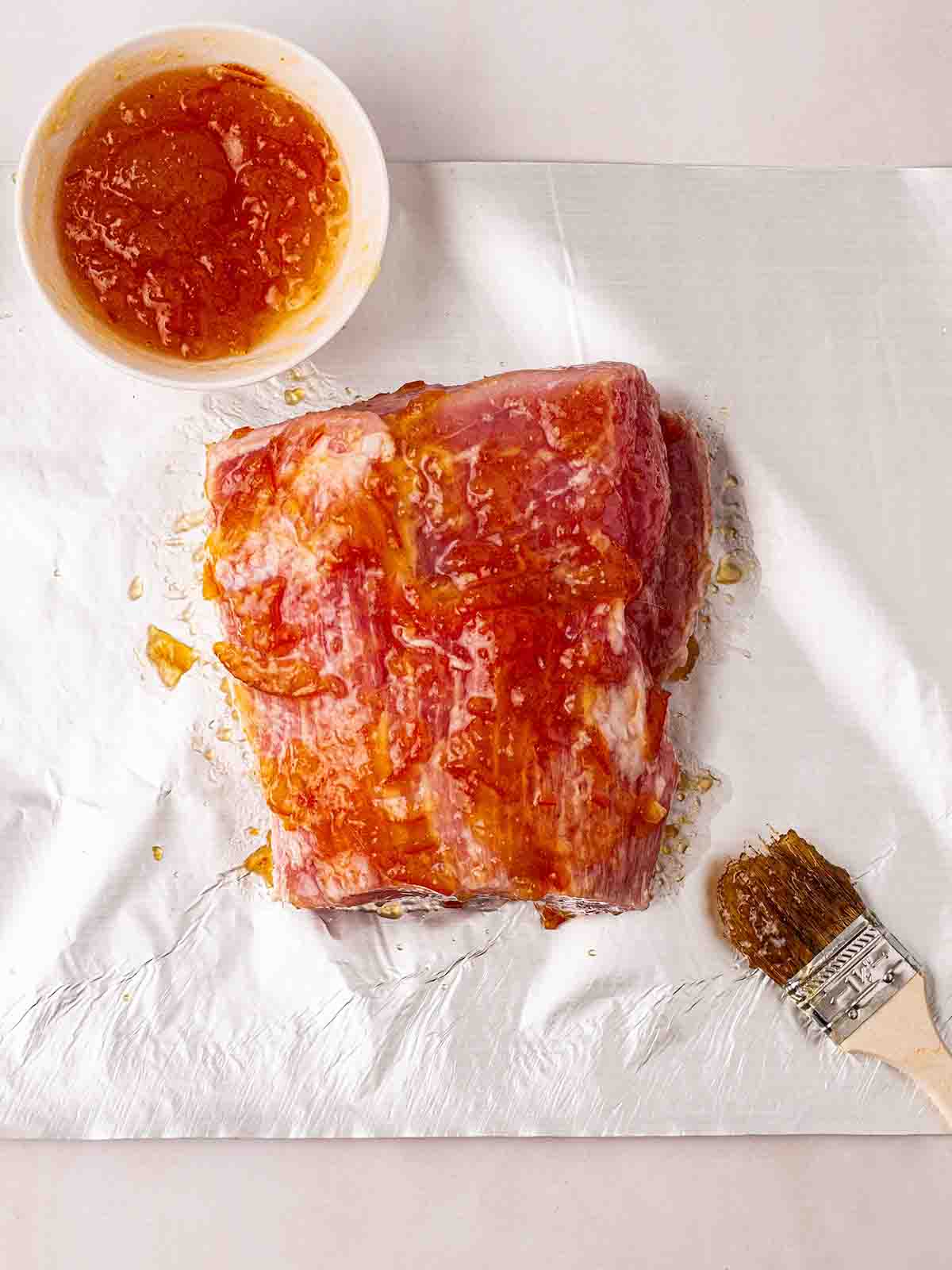 A gammon joint with a manmade and honey glaze brushed on top. It sits on a case of foil and the glaze is in a bowl to the side, with a brush that's been used to glaze.