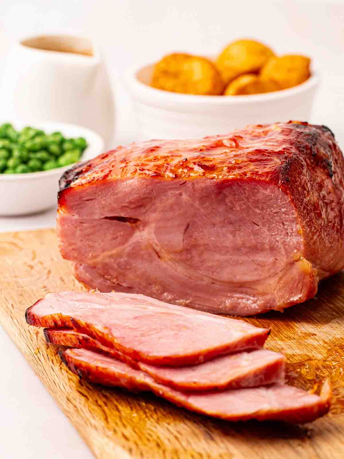 A cooked joint of gammon, having been cooked in an air fryer, on a wooden chopping board, sliced and ready to serve with peas and potatoes in the background.