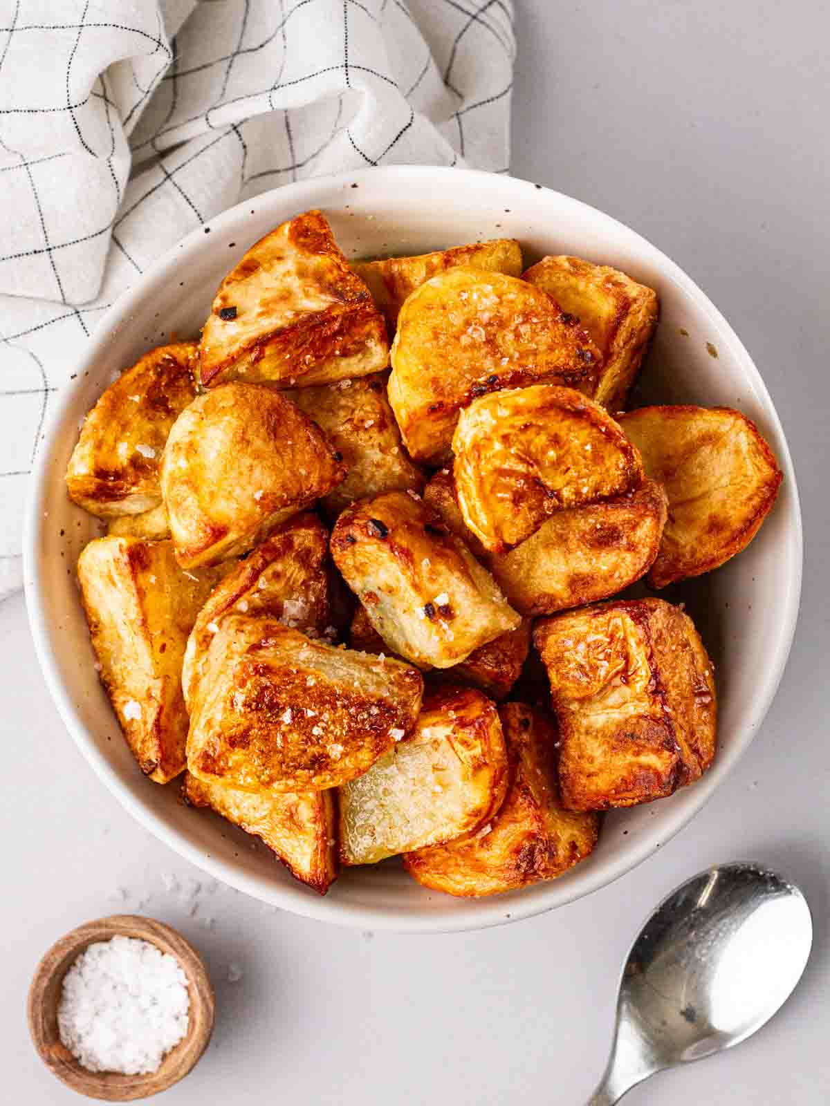 A bowlful of roast potatoes, cooked in the air fryer, on a white counter and surrounded by a spoon, salt pot and white cloth.
