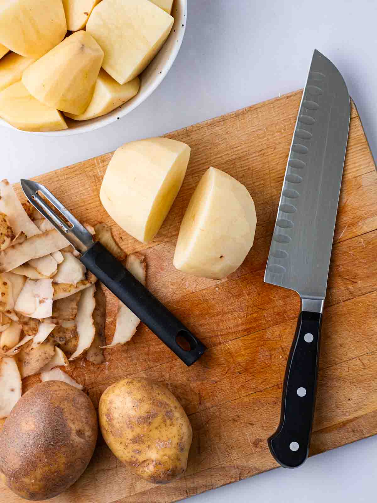 A chopping board with a knife and peeler, peeled potatoes, peelings and unpeeled potatoes, for step 1 in the recipe for air fryer roast potatoes.