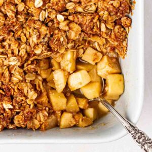 The corner of a pie dish with an Apple Crumble inside, with a spoonful missing, showing the apple filling and golden, crunchy topping, with a spoon sticking out.