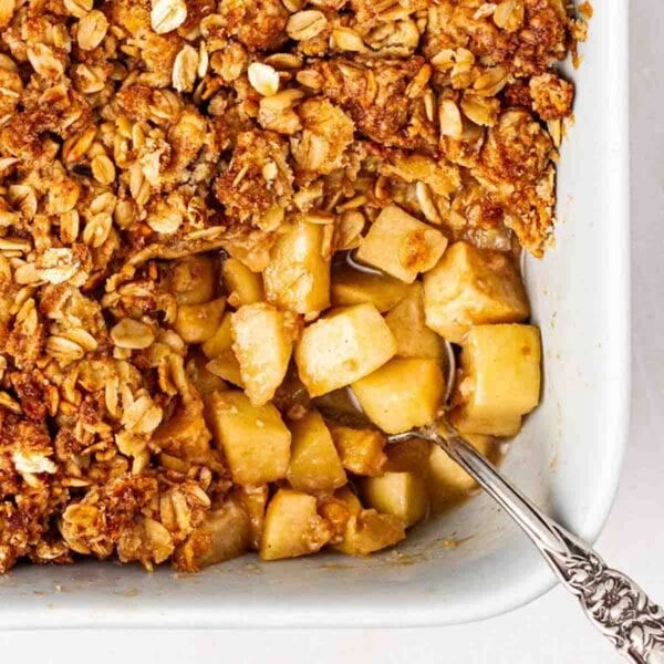 The corner of a pie dish with an Apple Crumble inside, with a spoonful missing, showing the apple filling and golden, crunchy topping, with a spoon sticking out.