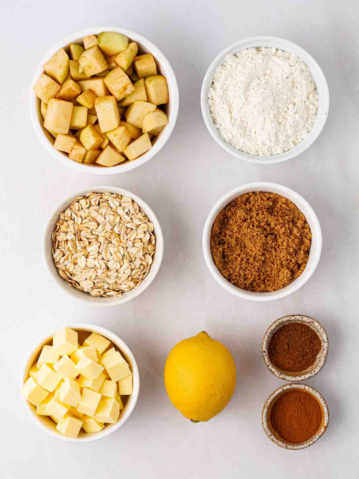 The ingredients for an Apple Crumble laid out in bowls on a white counter.