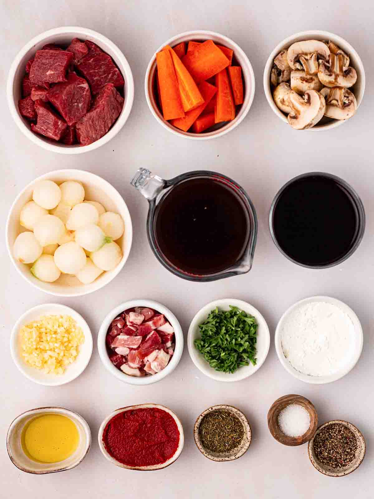 The ingredients for making beef bourguignon laid out in small bowls on a white counter top.