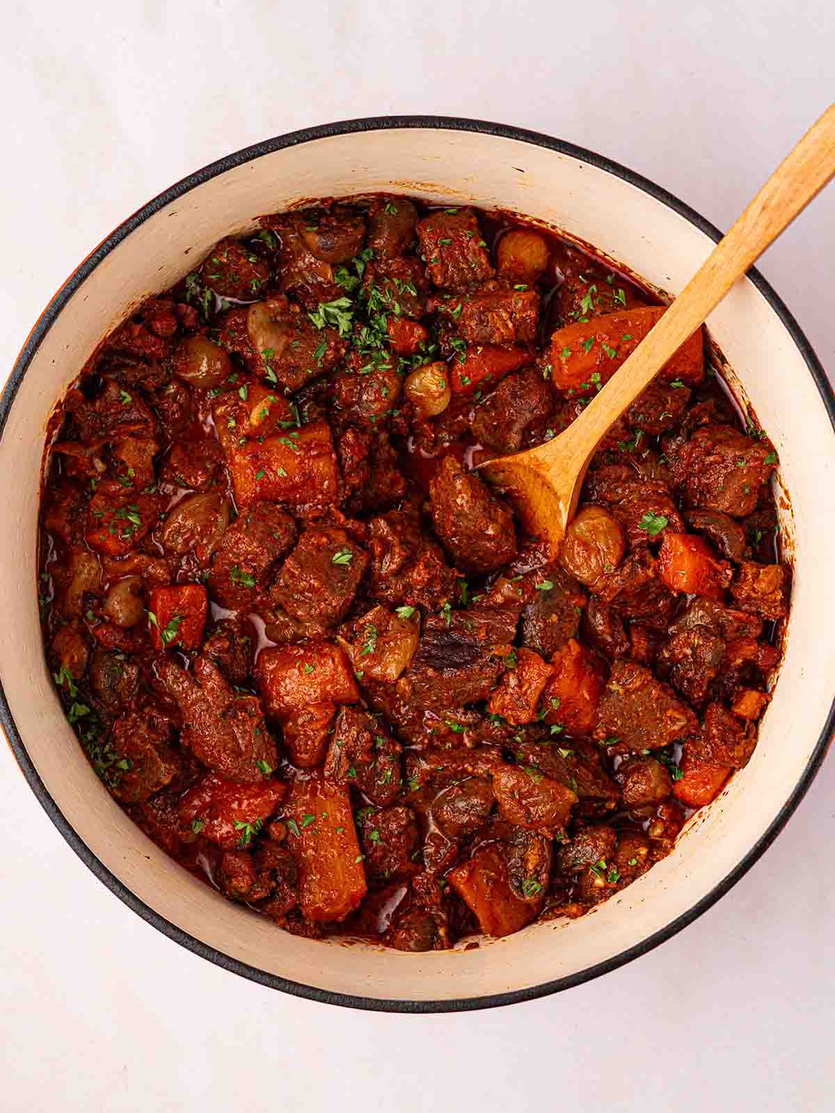 A pan filled with cooked beef bourguignon with a wooden spoon stirring, ready to serve.