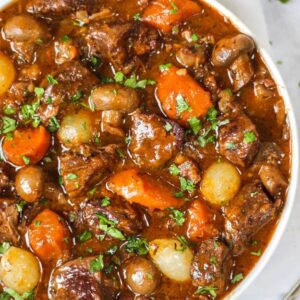 A big bowl of beef bourguignon on a white surface, ready to eat. Filled with meat, onions, carrots in a rich gravy.