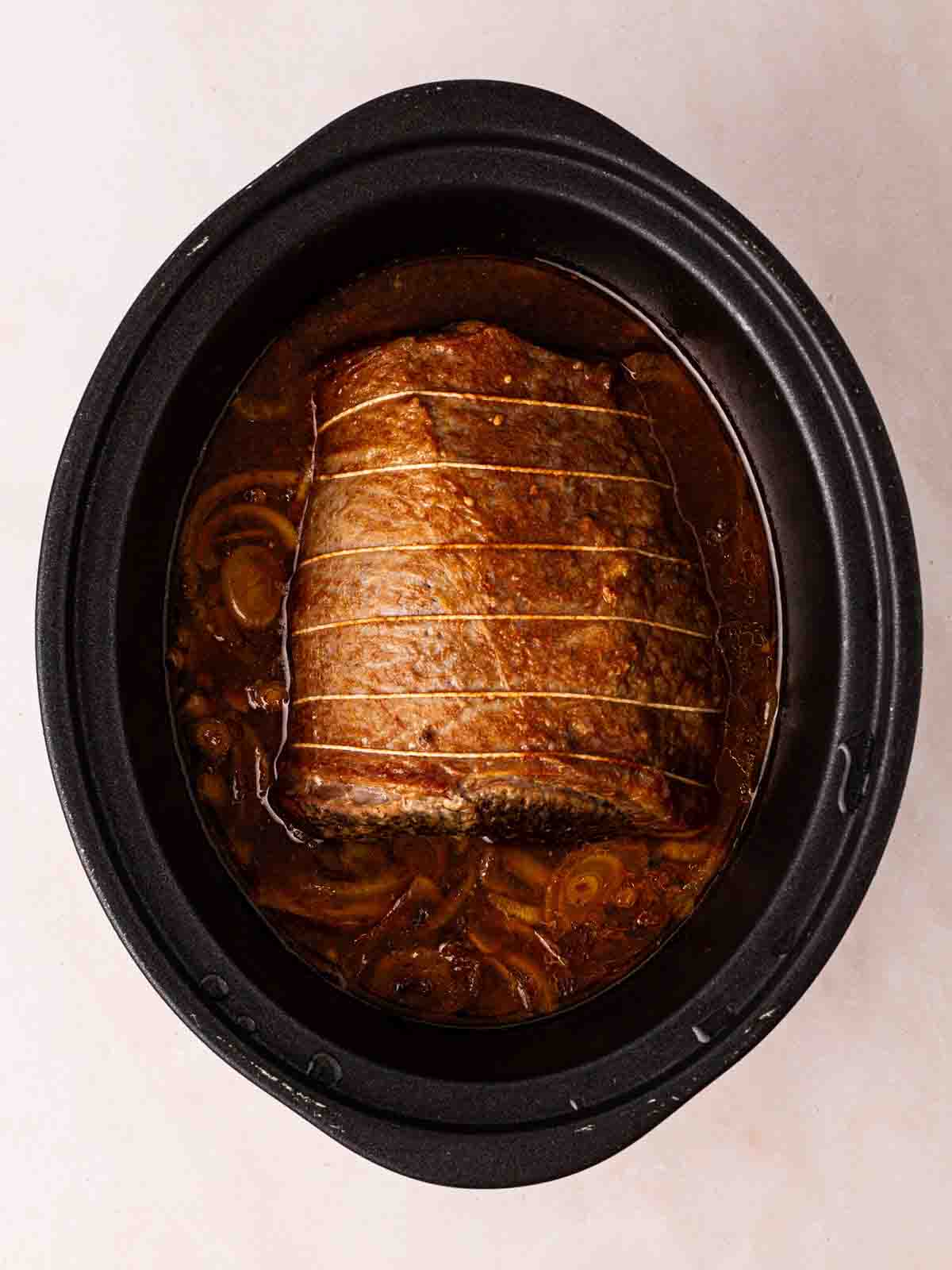 A cooked piece of beef brisket with onions in a slow cooker pan.
