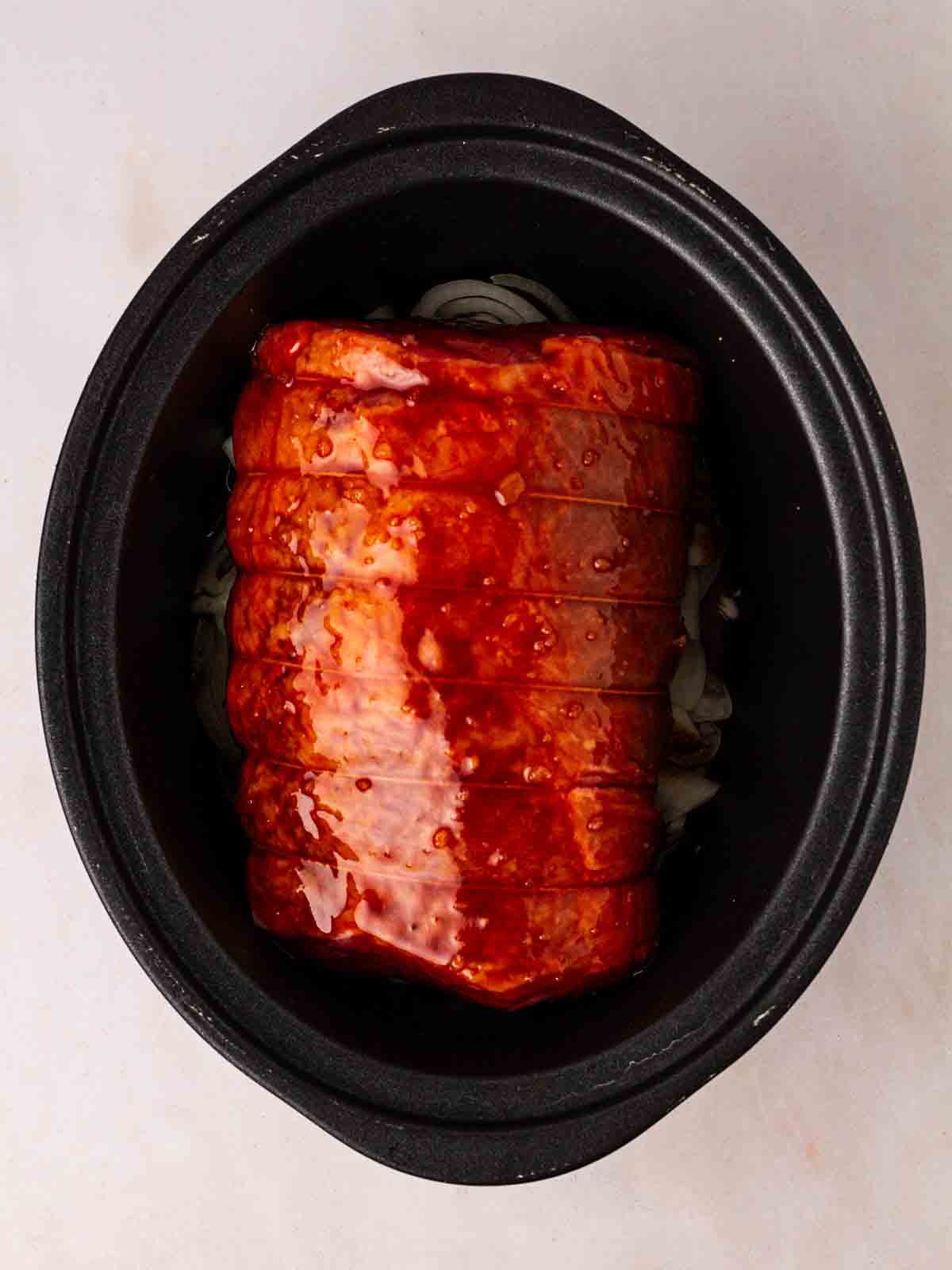 A piece of raw beef brisket in a slow cooker, topped with barbecue sauce, ready to be cooked.