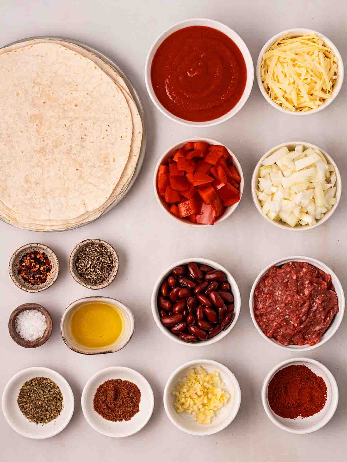The ingredients for making Beef Enchiladas laid out in bowls and plates on a white surface.
