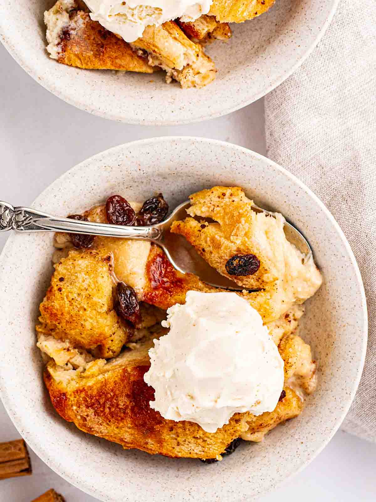 Two bowls, with one fully in shot, with bread and butter pudding inside, served with ice cream. A spoon sits in the bowl.