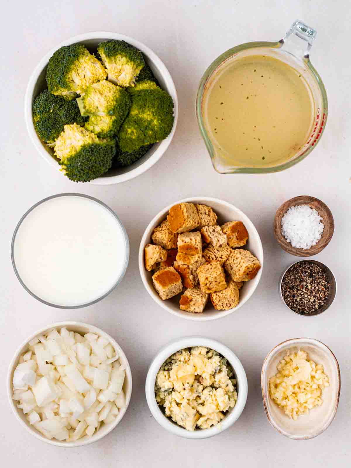 The ingredients for making Broccoli and Stilton Soup laid out in bowls on a white counter top.