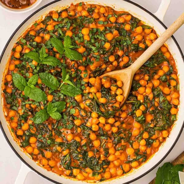 A large white pan filled with Chickpea Curry with spinach and mint garnish, and a wooden spoon stirring.