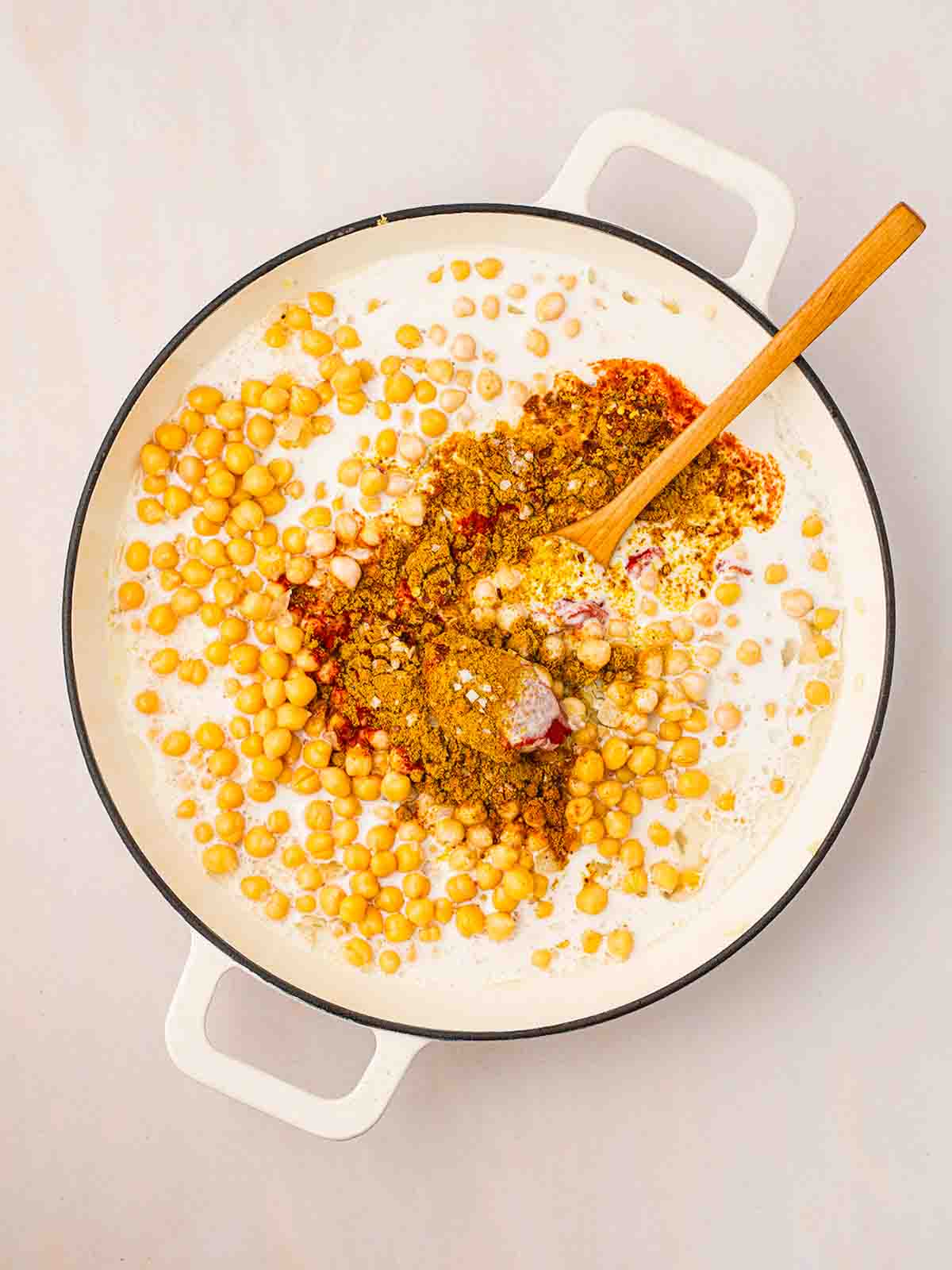A pan filled with uncooked chickepea curry ingredients, being stirred with a wooden spoon, for step 2 in the process for Chickpea Curry recipe.