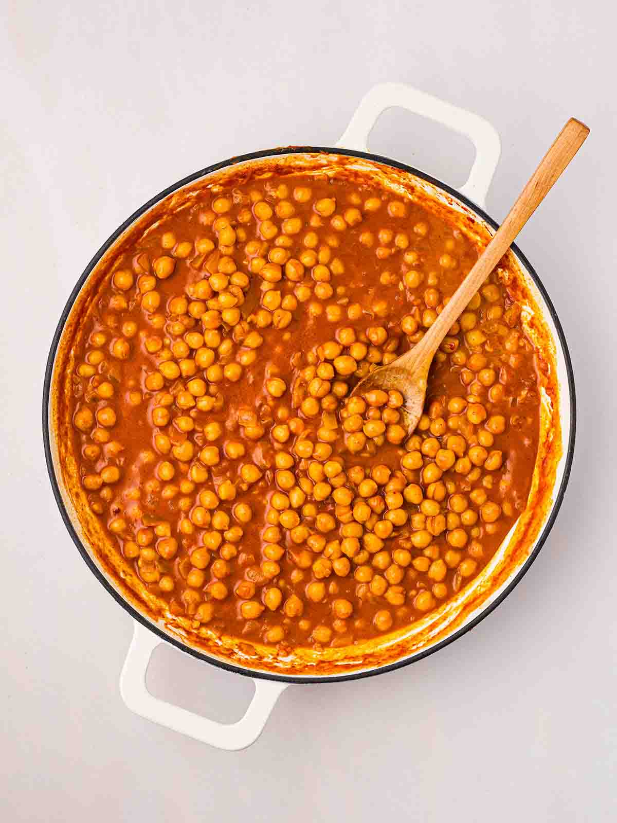 Chickpeas in a curry sauce in pan, being stirred with a wooden spoon, for step 3 in the recipe for Chickpea Curry.