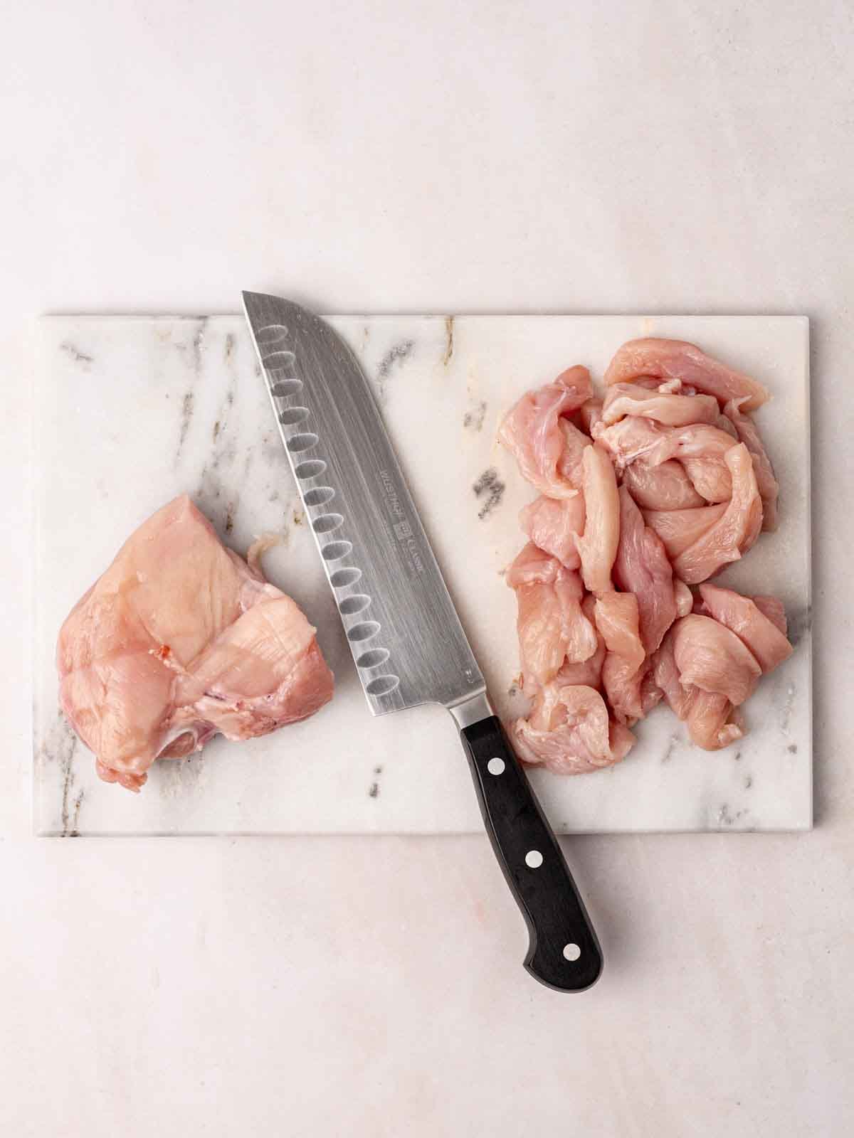 A marble chopping board with a knife and raw chicken breast, with half already cut into bitesized pieces for making Chicken Goujons.
