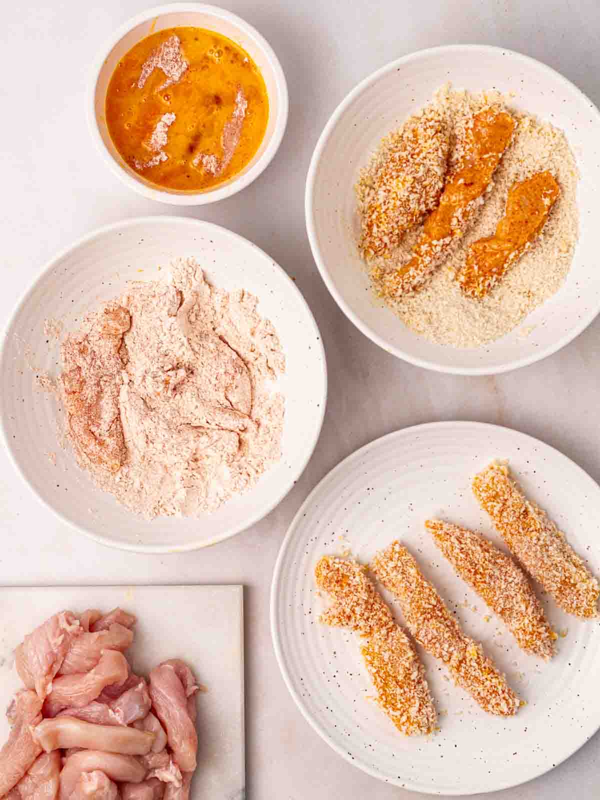 Several bowls of ingredients to coat pieces of raw chicken in breadcrumbs to make homemade Chicken Goujons.