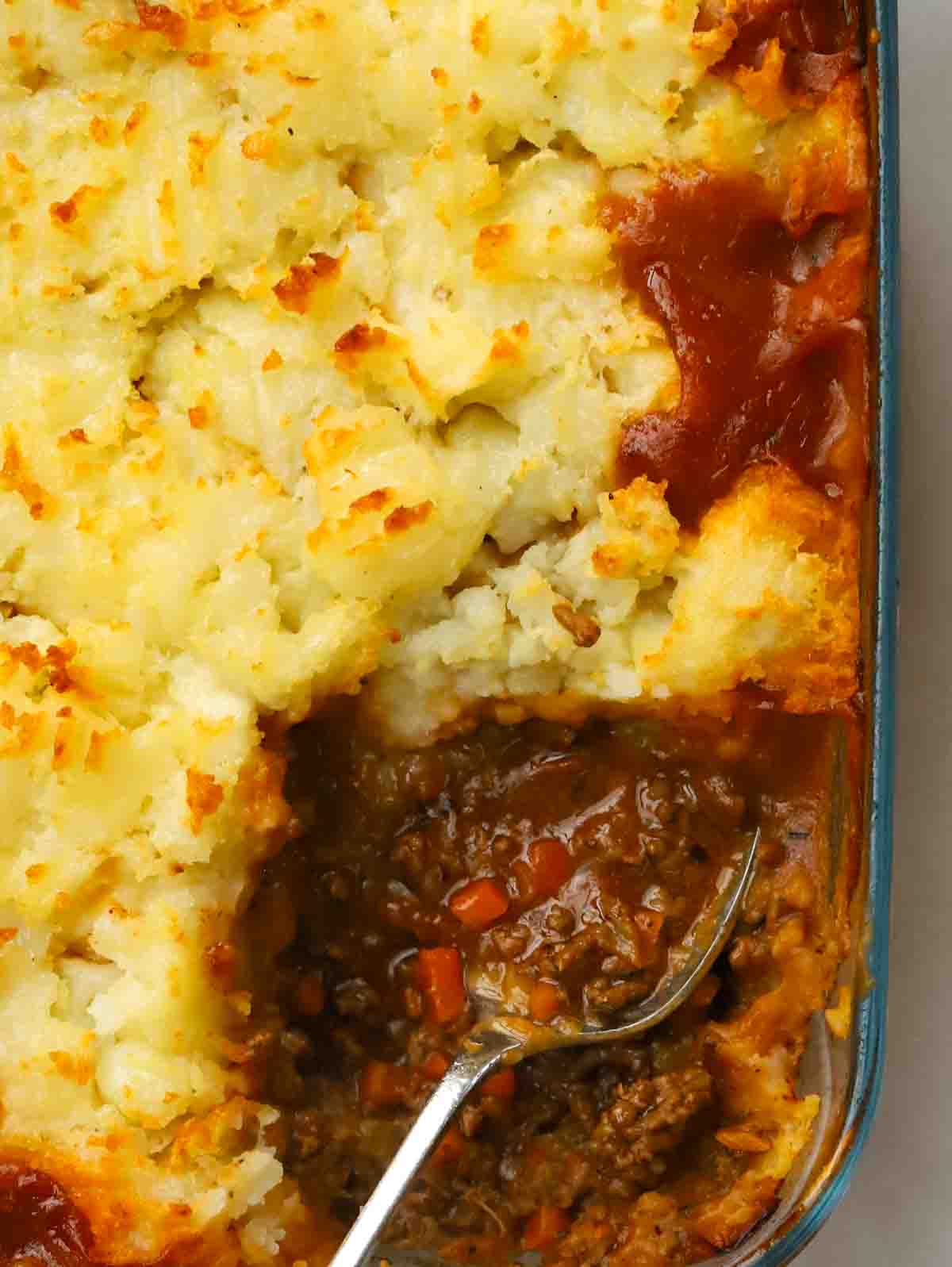 A close up of a cottage pie with one corner missing after being served, to reveal the inside of the pie, with a spoon dishing out.