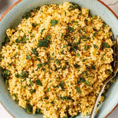 A big bowl of fluffy couscous with a fork in, ready to eat, on a table with a small bowl of salt and mint, and a tea towel.
