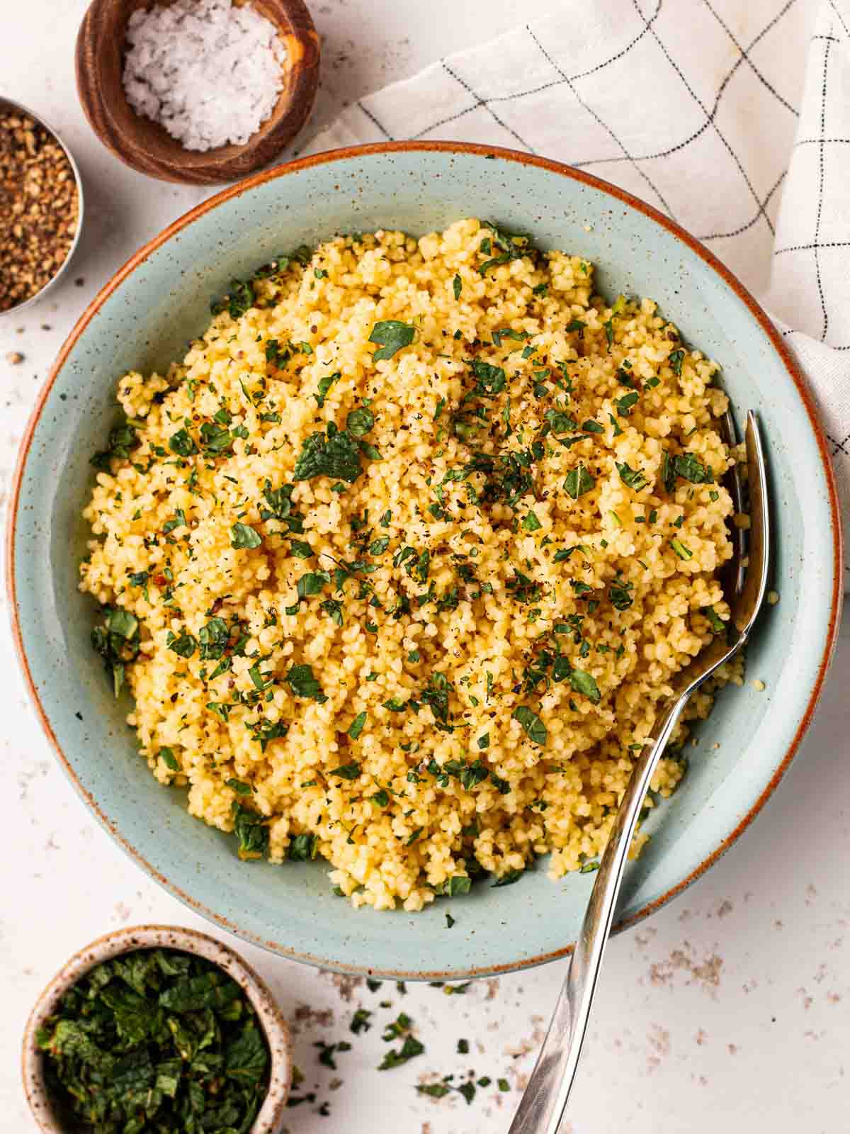 A blue bowl filled with couscous with a fork stuck in, surrounded by small pinch pot bowls of salt and mint.