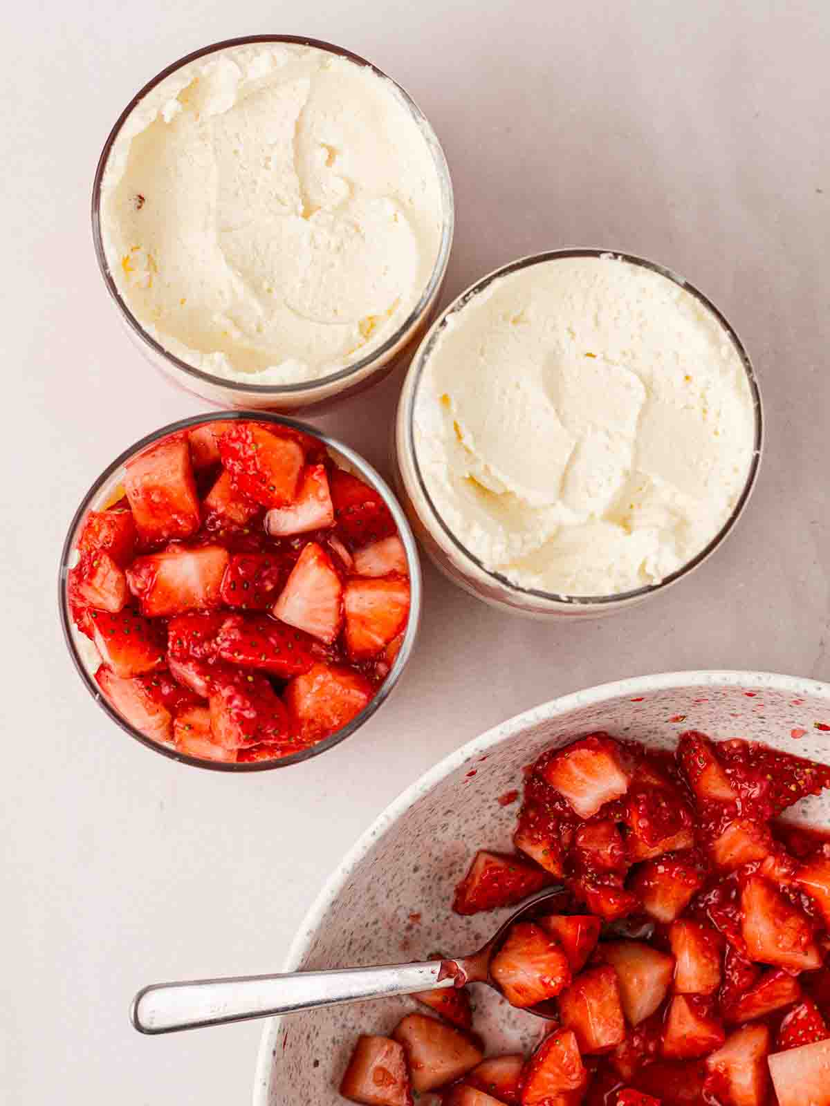 A bowl filled with strawberries and glasses being layered with cream and meringue for individual Eton Mess desserts.