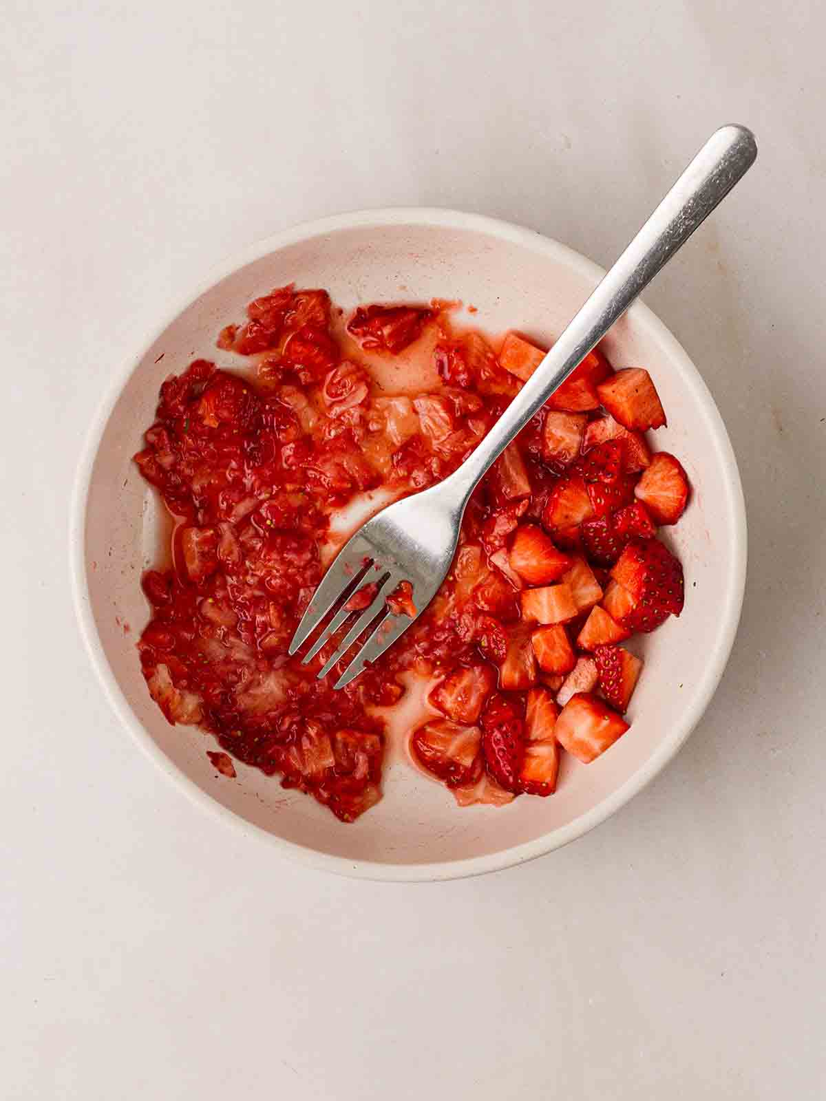 A white bowl with strawberries being mushed up with a fork for step 3 in the recipe for Eton Mess.