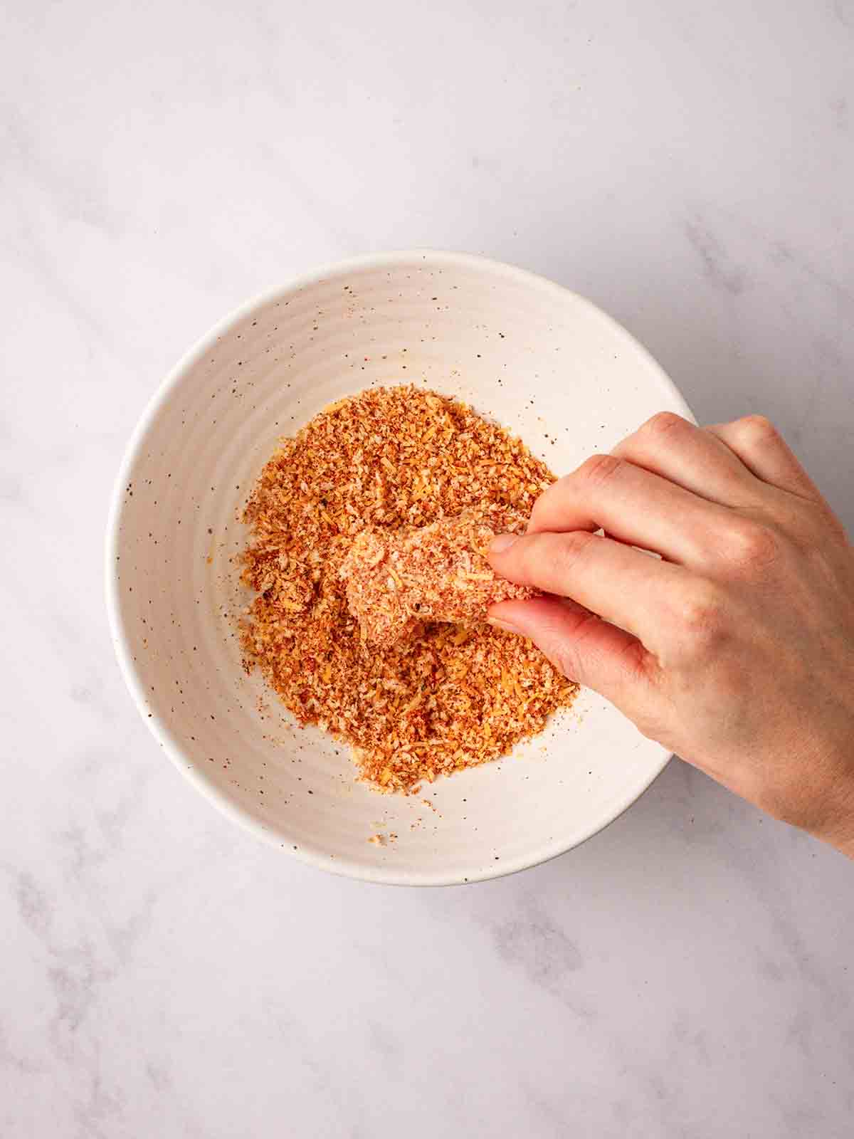 A white bowl with breadcrumb ingredients, with a hand dipping in a piece of raw chicken for the recipe homemade chicken nuggets.