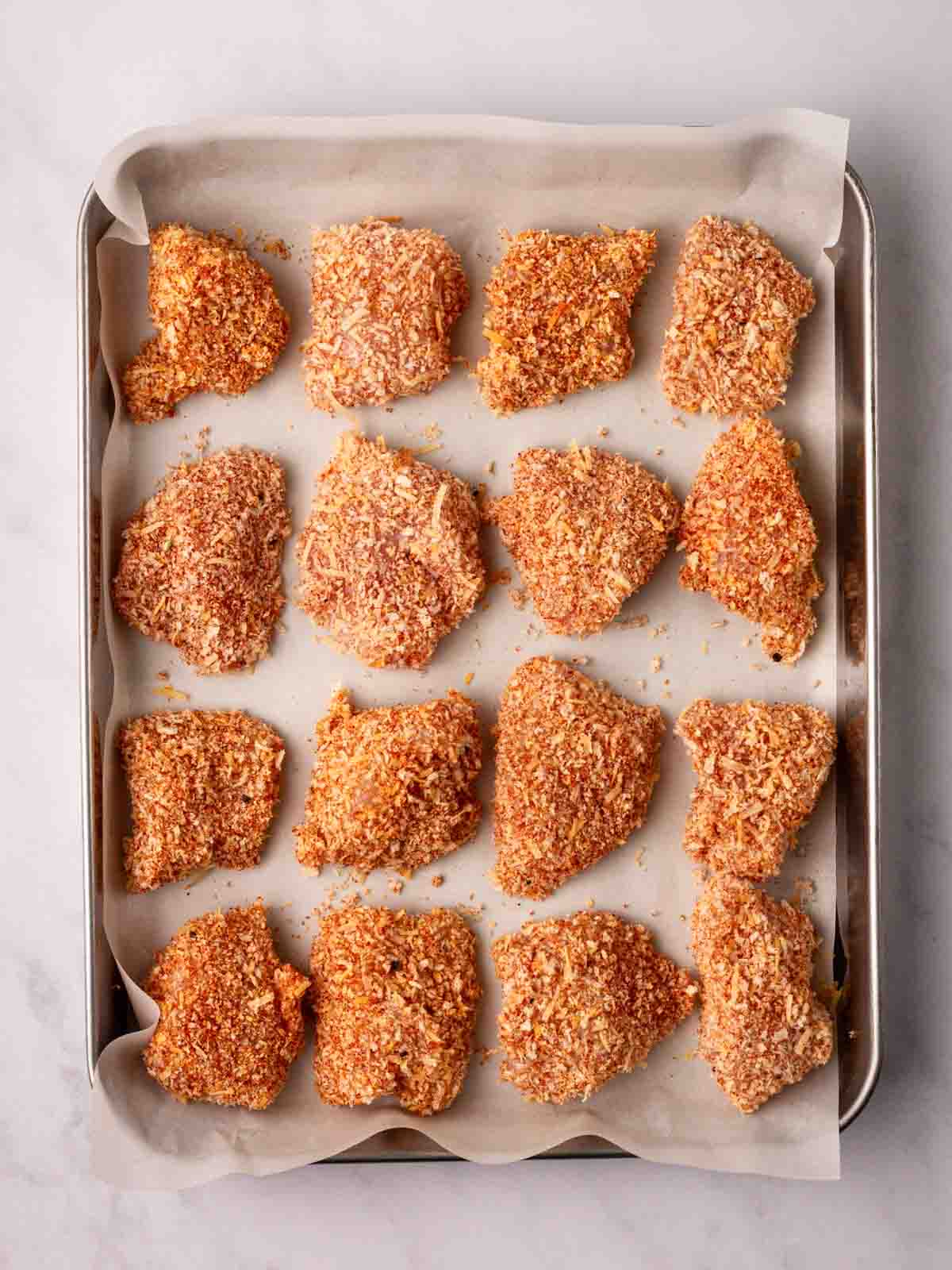 Pieces of coated, raw homemade chicken nuggets on a lined baking tray, ready to cook.