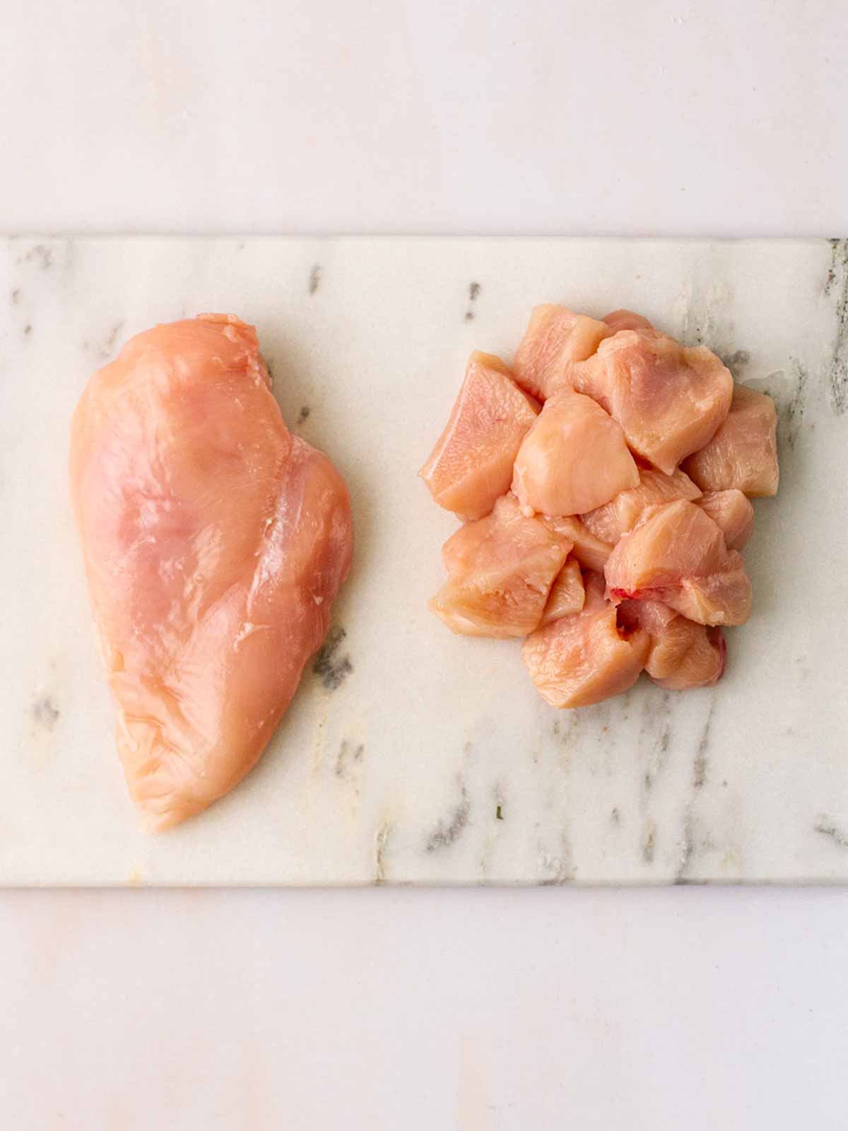 A chicken breast on the left hand side of a board, and cubes of chicken breast on the right hand side to show what it looks like chopped into pieces.