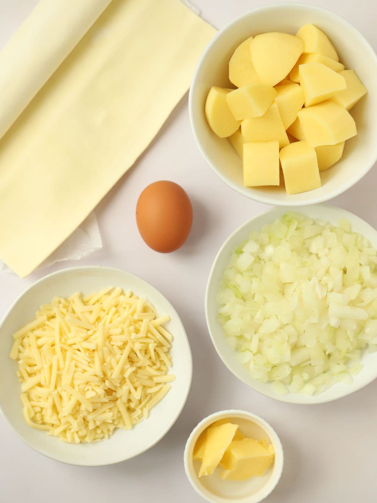 The ingredients for homemade Cheese and Onion Pasties laid out on a counter top in bowls. Includes Pastry, potatoes, and egg, cheese, chopped onion and butter.