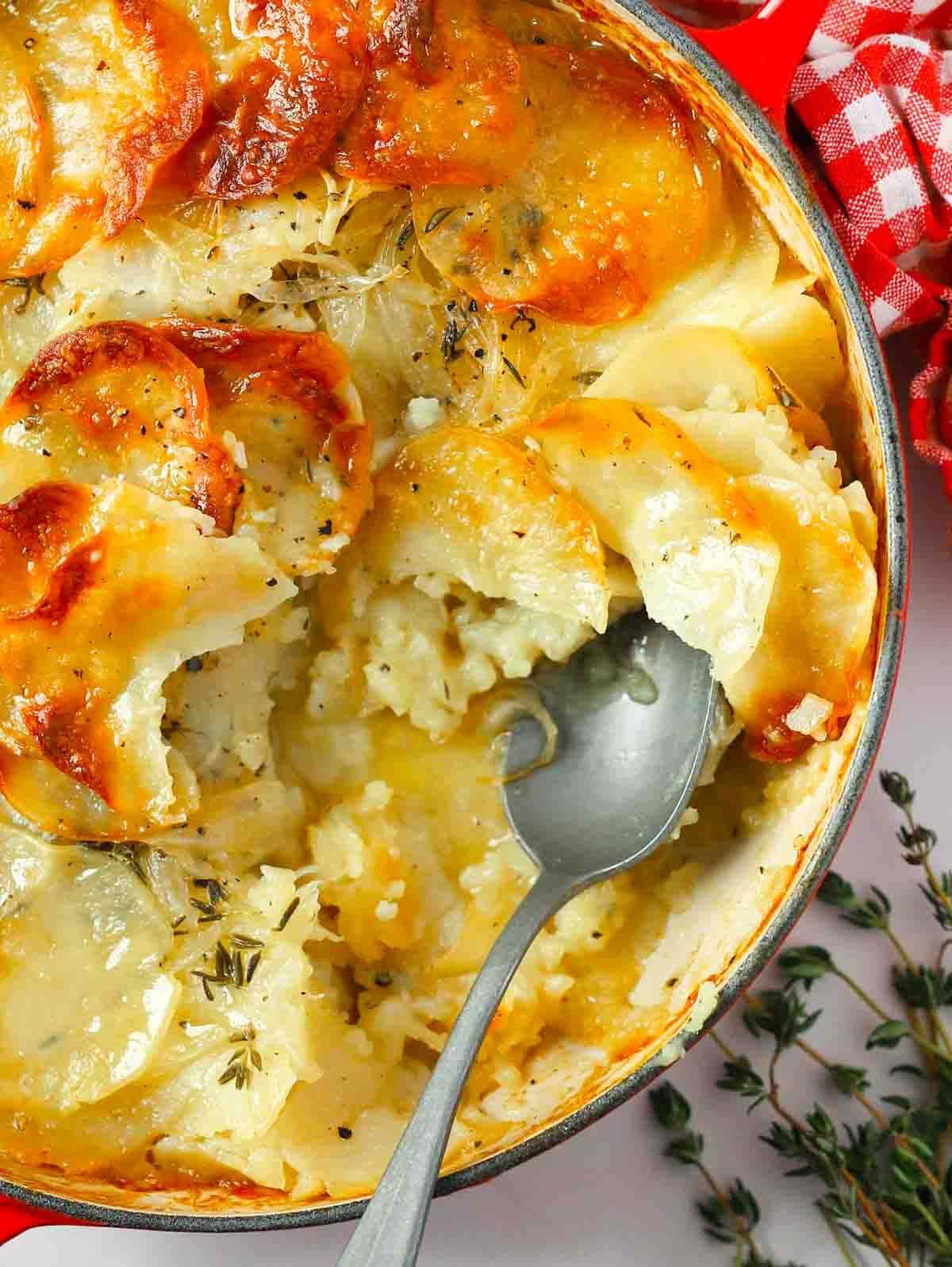 A pan filled with cooked Boulangere Potatoes, with a spoon in and one portion missing.