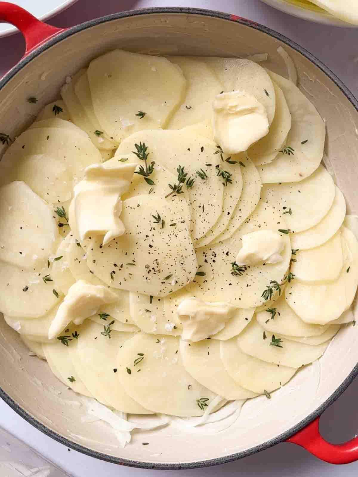 Slices of raw potatoes in a big pan, with butter and herbs on top for step 3 in the recipe for Boulangere Potatoes.
