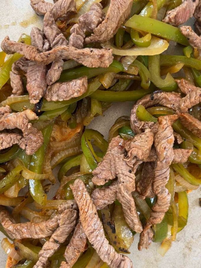 Strips of cooked steak and peppers and onions on top of an open baguette for step 3 in the recipe for Philly Cheesesteak.