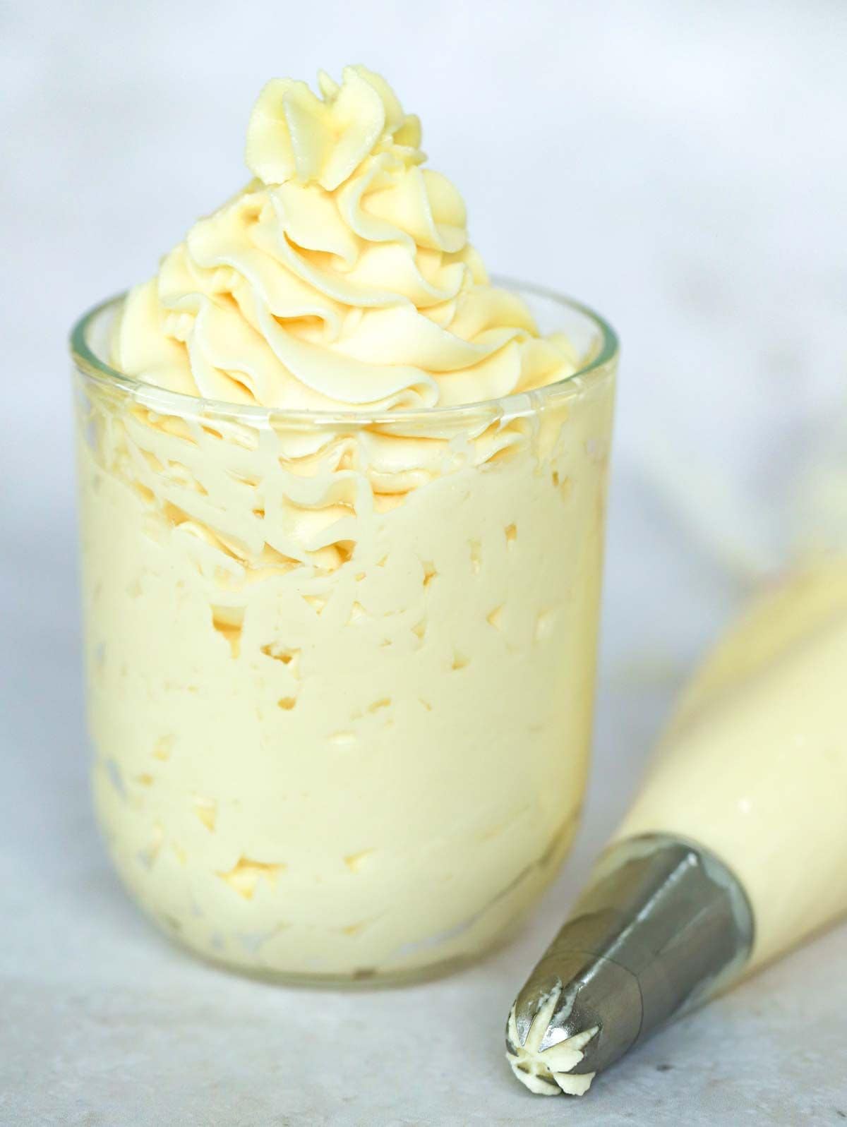 A small glass filled with cream cheese frosting, with a full icing bag laid at the side of it.