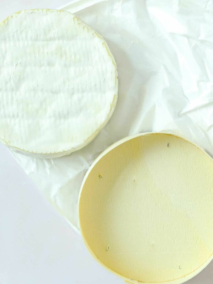 A camembert wheel next to its wooden box, on top of its wrapper, on a white surface.