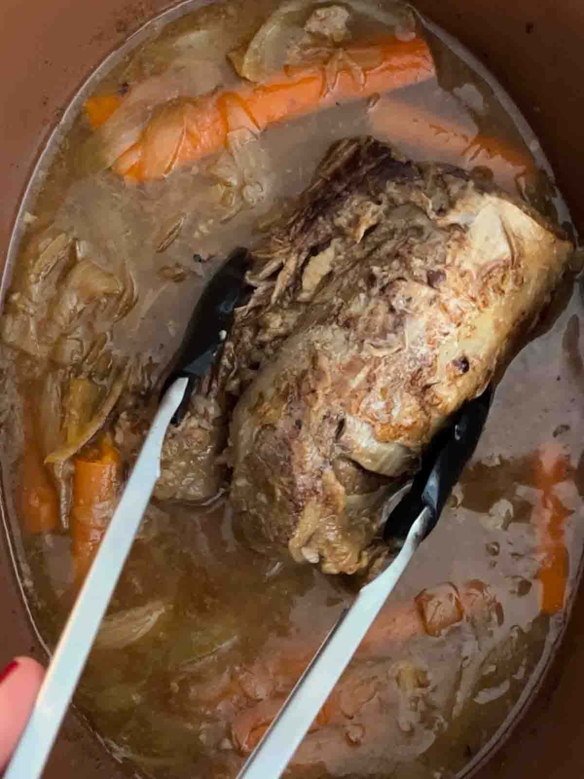 A slow cooker joint of beef in gravy with tong around it, about to bring it out.