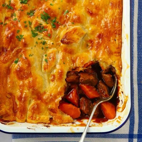 The corner of a puff pastry steak pie with a corner missing, with a spoon dipped in ready to serve a portion.