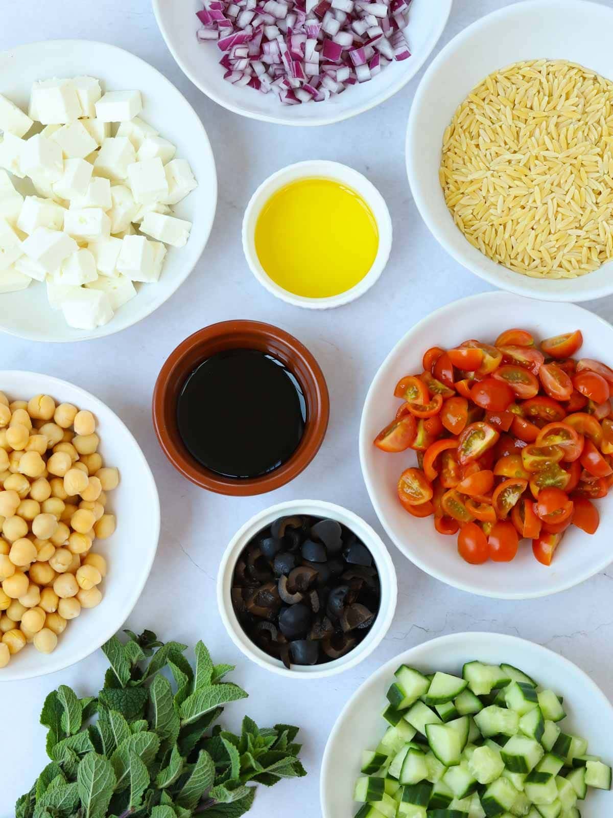Ingredients for the recipe Orzo Pasta Salad laid out on a counter. Onion, feta, orzo, tomatoes, chickpeas, cucumber, mind and dressing.