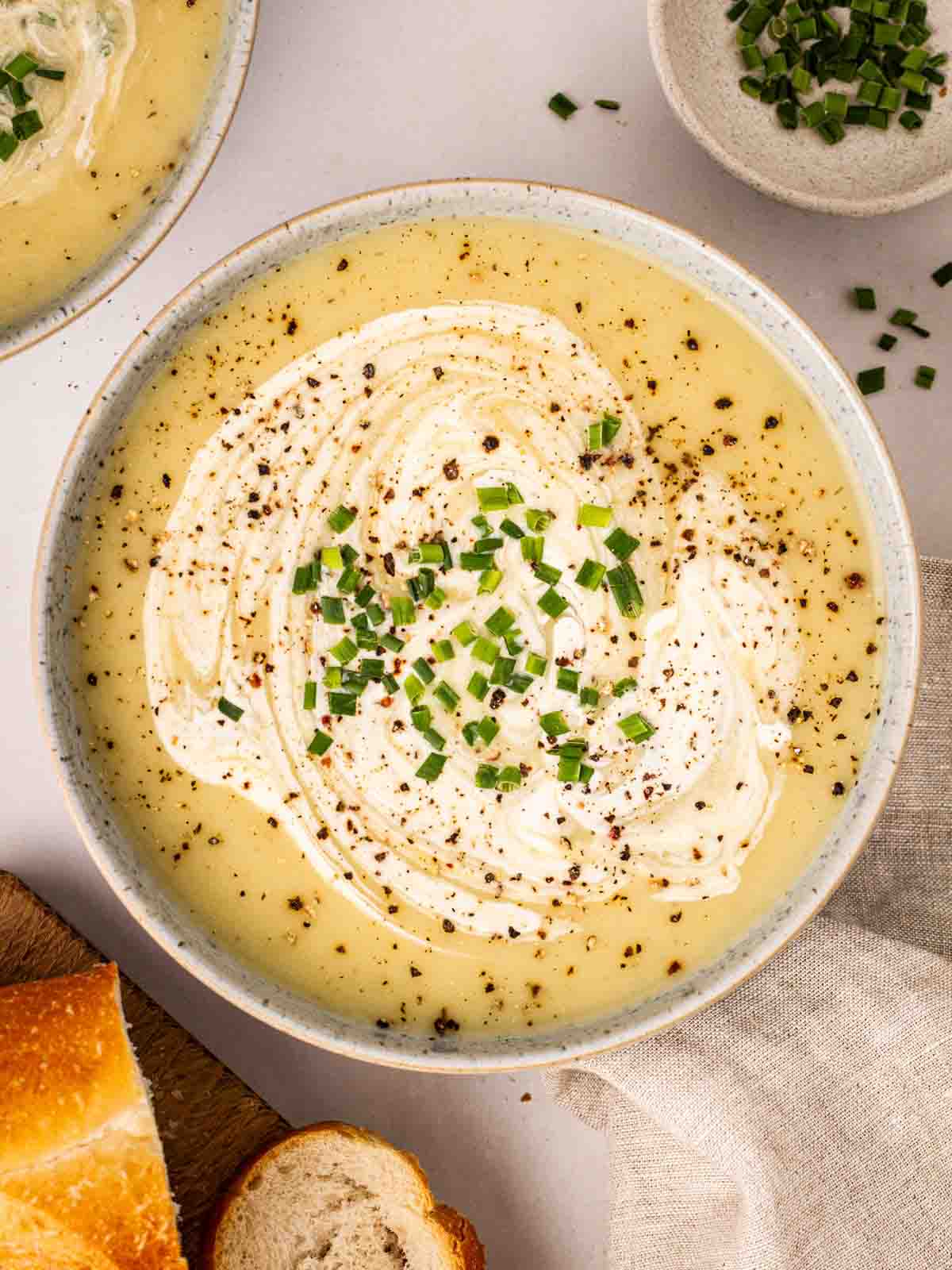 A bowl of creamy leek and potato soup on a table with surrounding bread and chives.