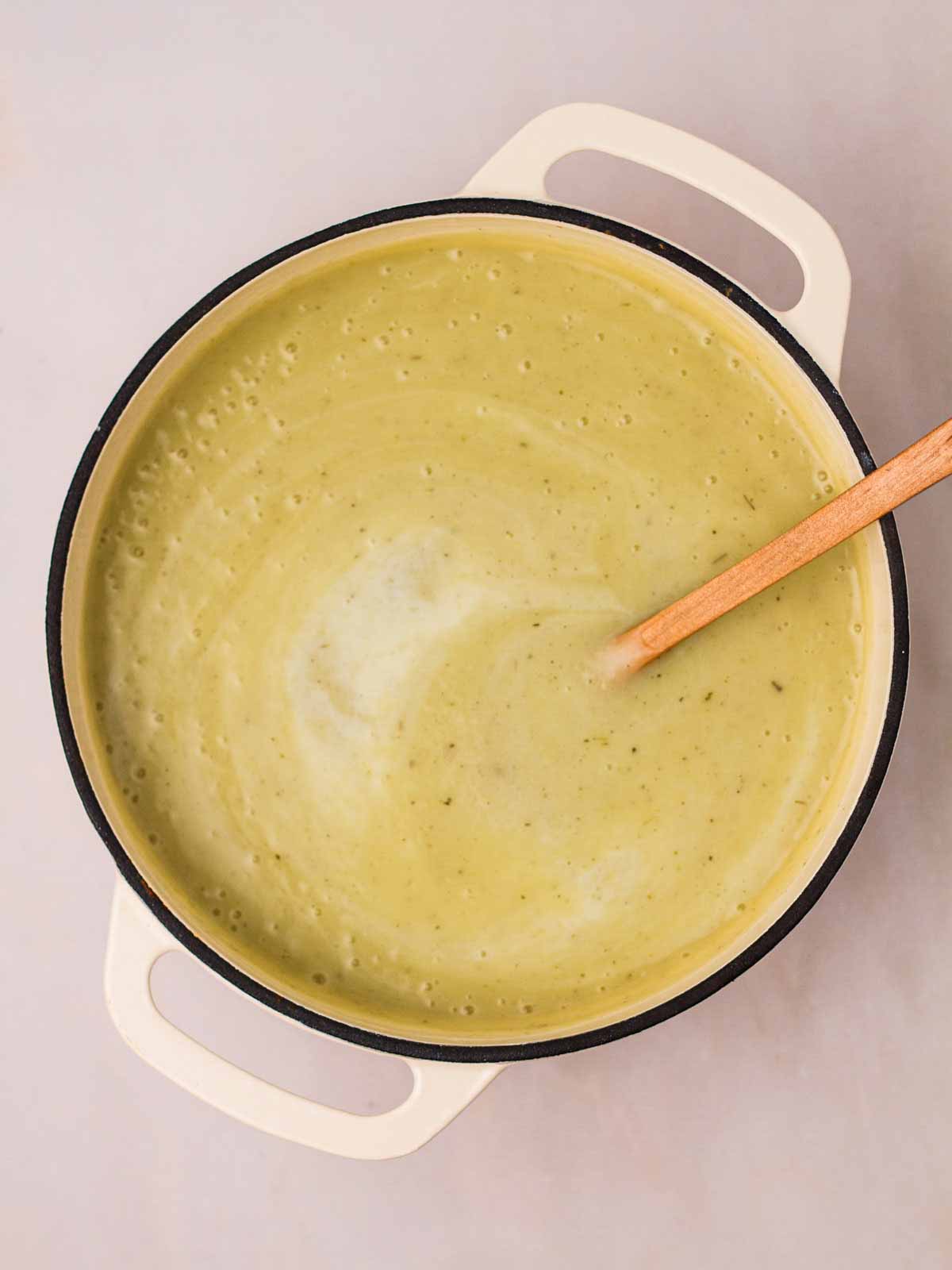 A smooth Leek and Potato Soup in a large pan with a wooden spoon sticking out, stirring in cream.