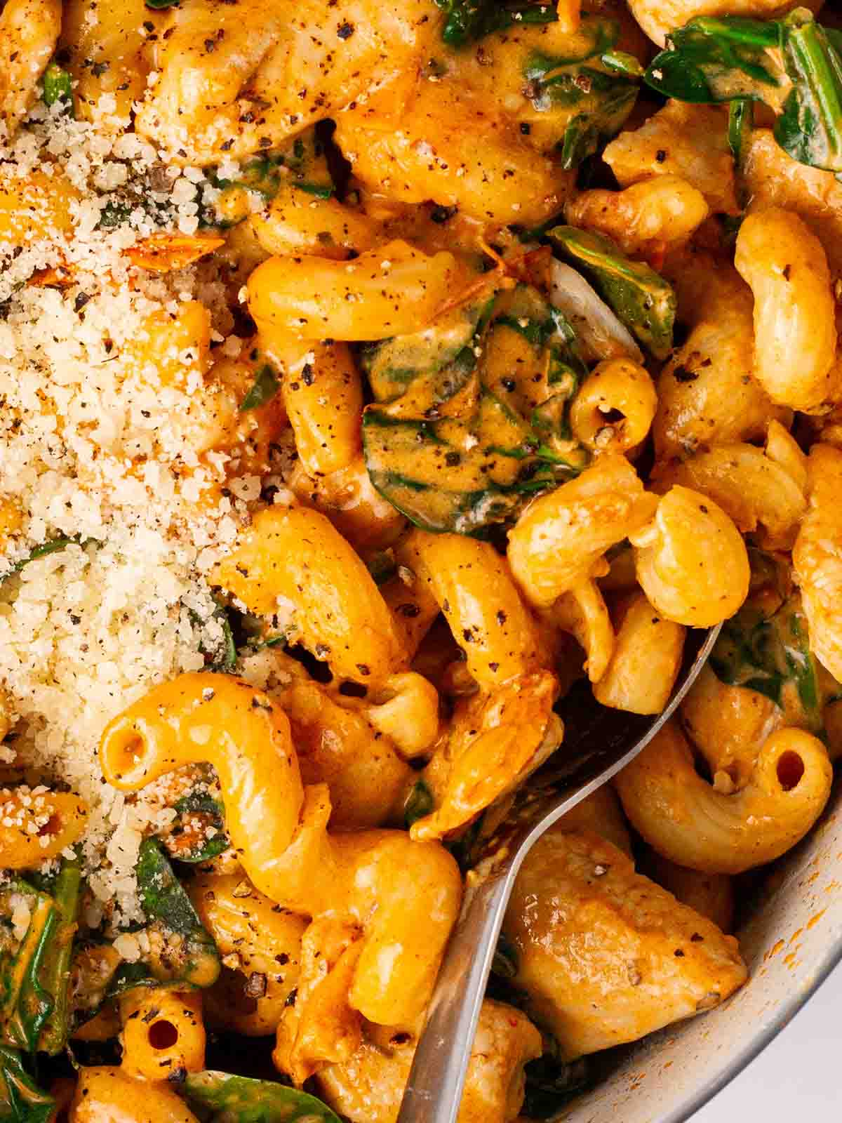 A close up of pasta in a pan with a spoon and cheese and spinach.