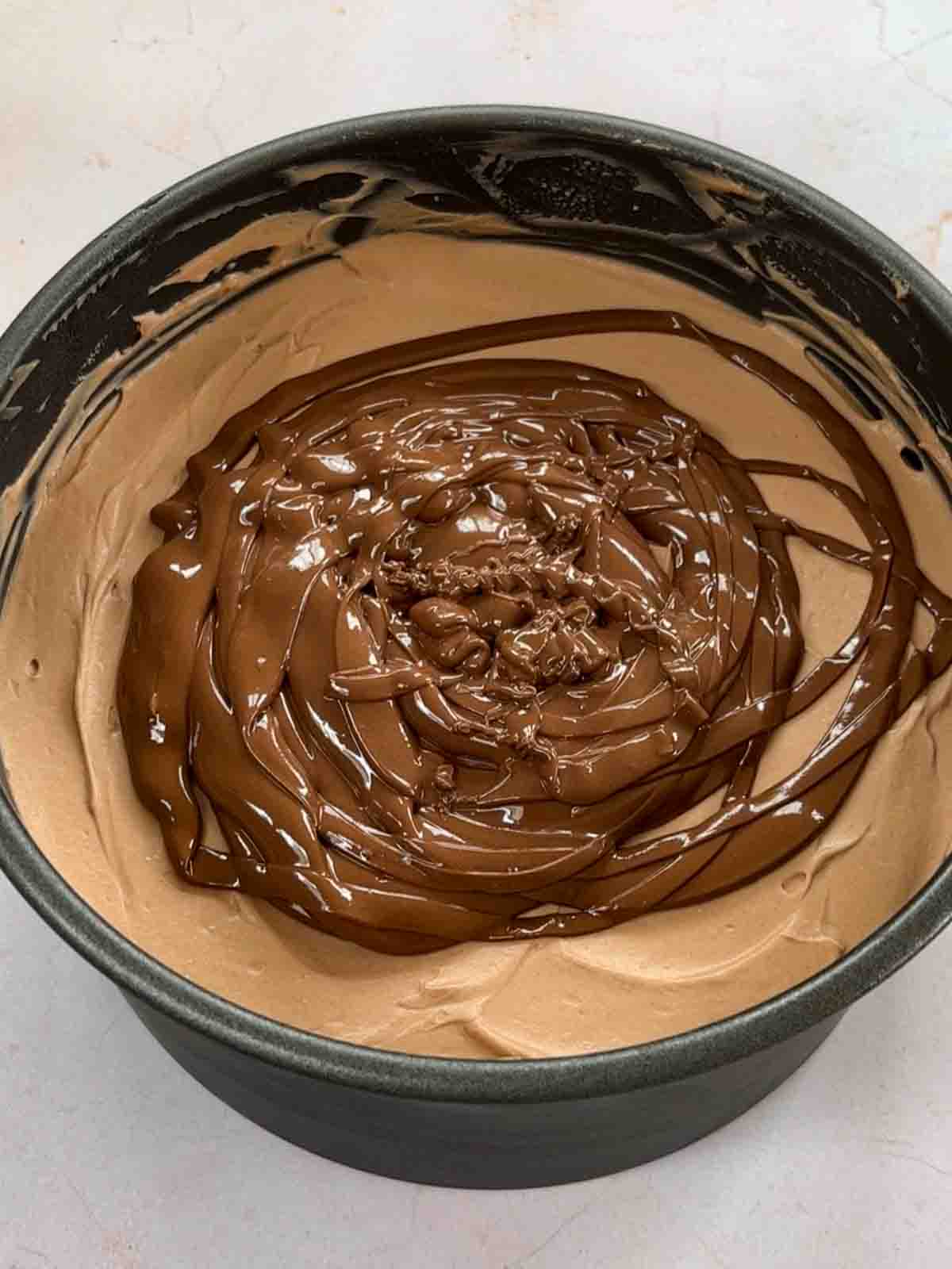 A baking tin filled with whipped cream mixture and topped with chocolate ready to spread for for step 5 in the recipe for Nutella Cheesecake.