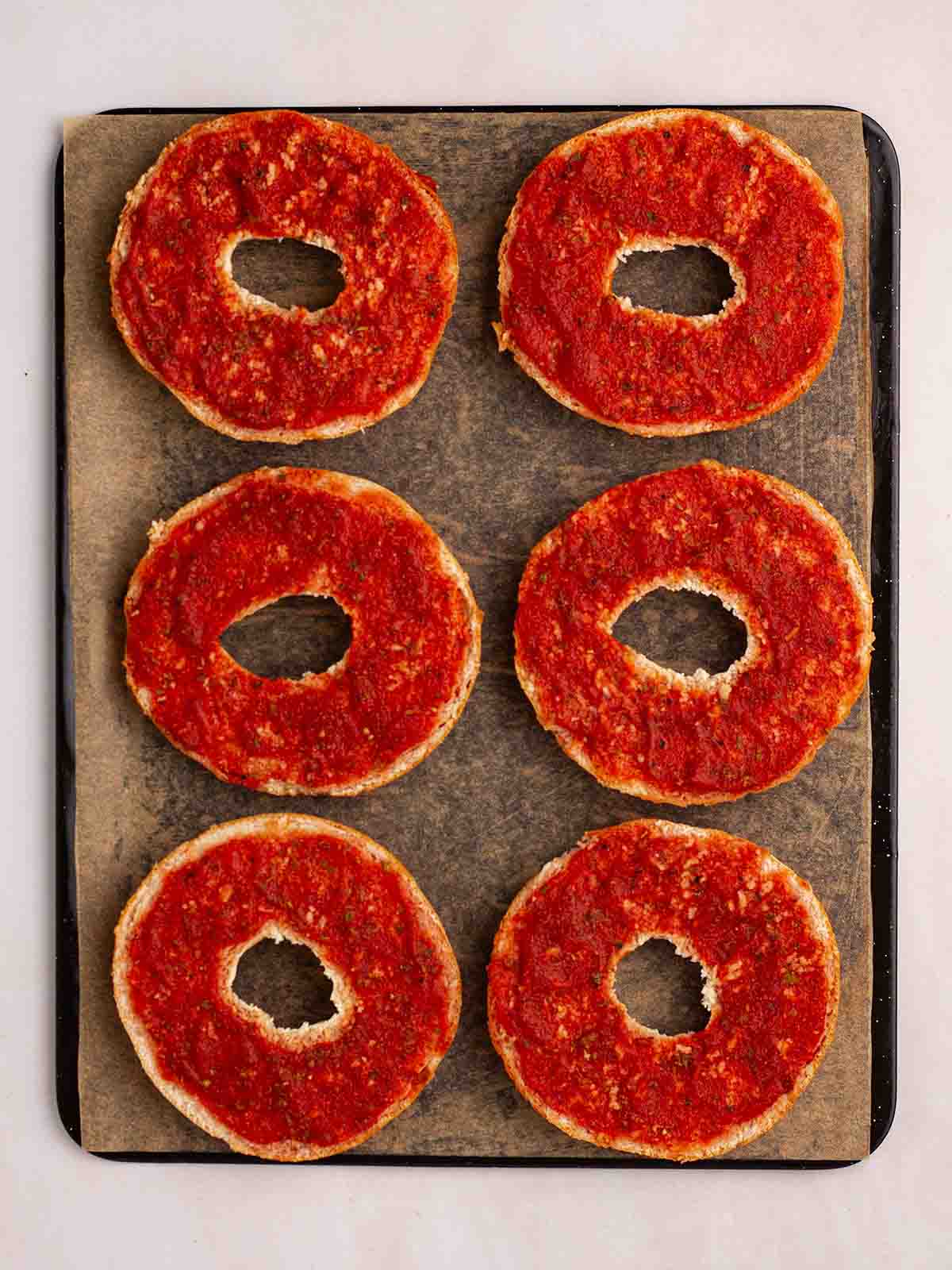 A baking tray with halved bagels with passata on top for step 2 in the recipe for Pizza Bagels.