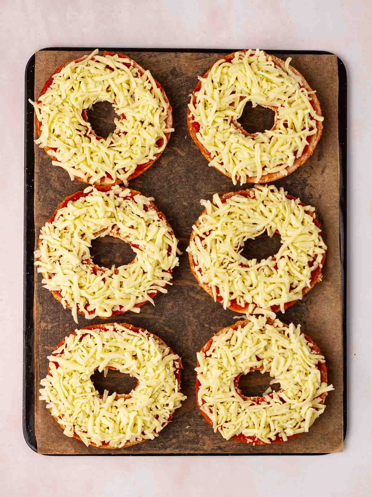 An oven tray with halved bagels with tomato sauce and cheese on top, ready to be cooked into pizza style bagels.
