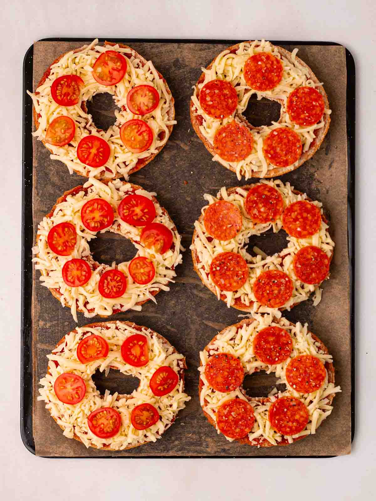 A baking tray with halved bagels on, topped with pepperoni and tomato, for step 4 in the recipe for Pizza Bagels.