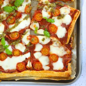 A baking tray with a baked Puff Pastry Pizza on top, with mozzarella, cherry tomatoes, basil and pesto as toppings.