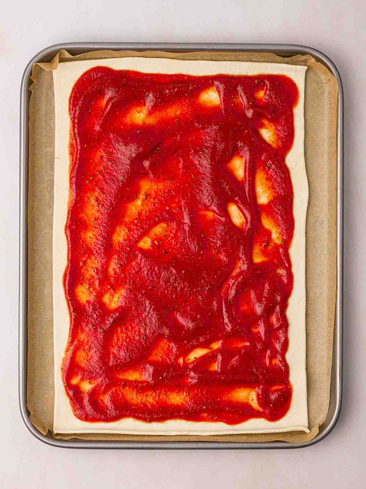 A baking tray with baking paper and a sheet of puff pastry on top, which is covered with tomato sauce for step 1 in the recipe for Puff Pastry Pizza.