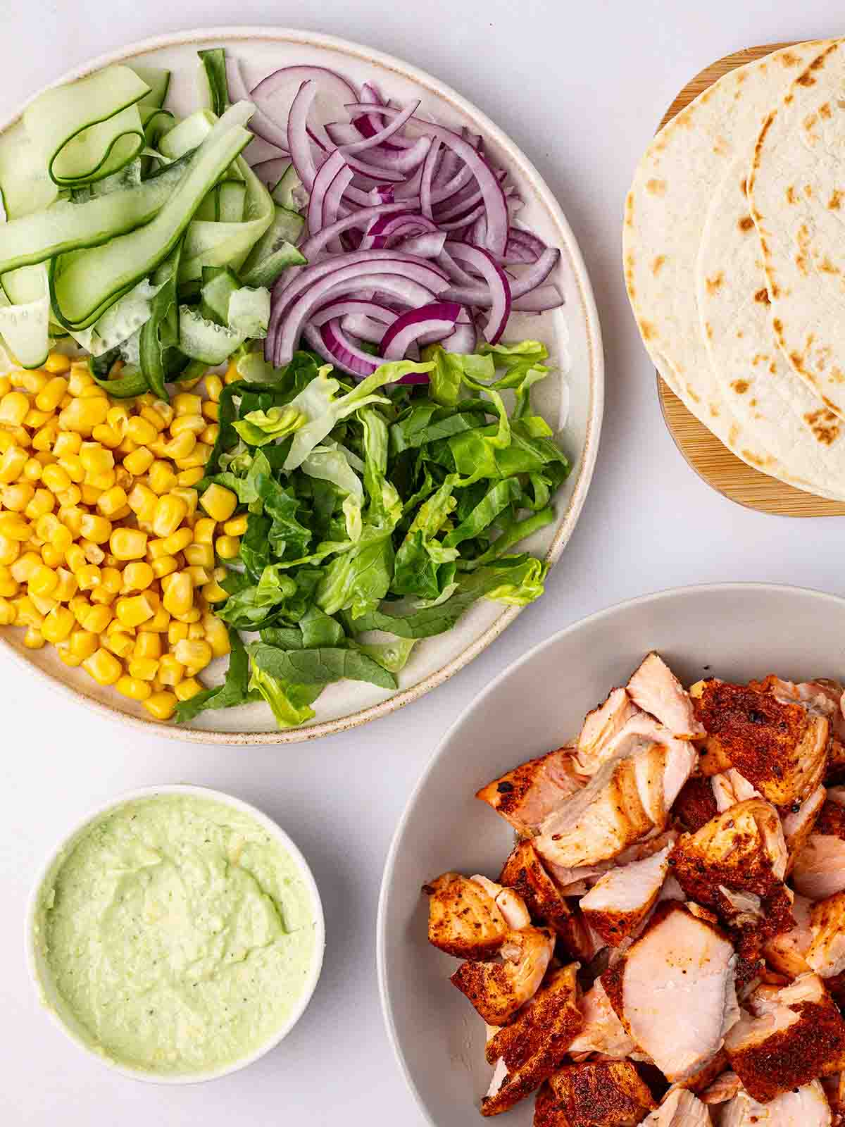 A bowl of salmon, a plate of salad, a pot of avocado dressing and a plate of wraps to make up Salmon Tacos.
