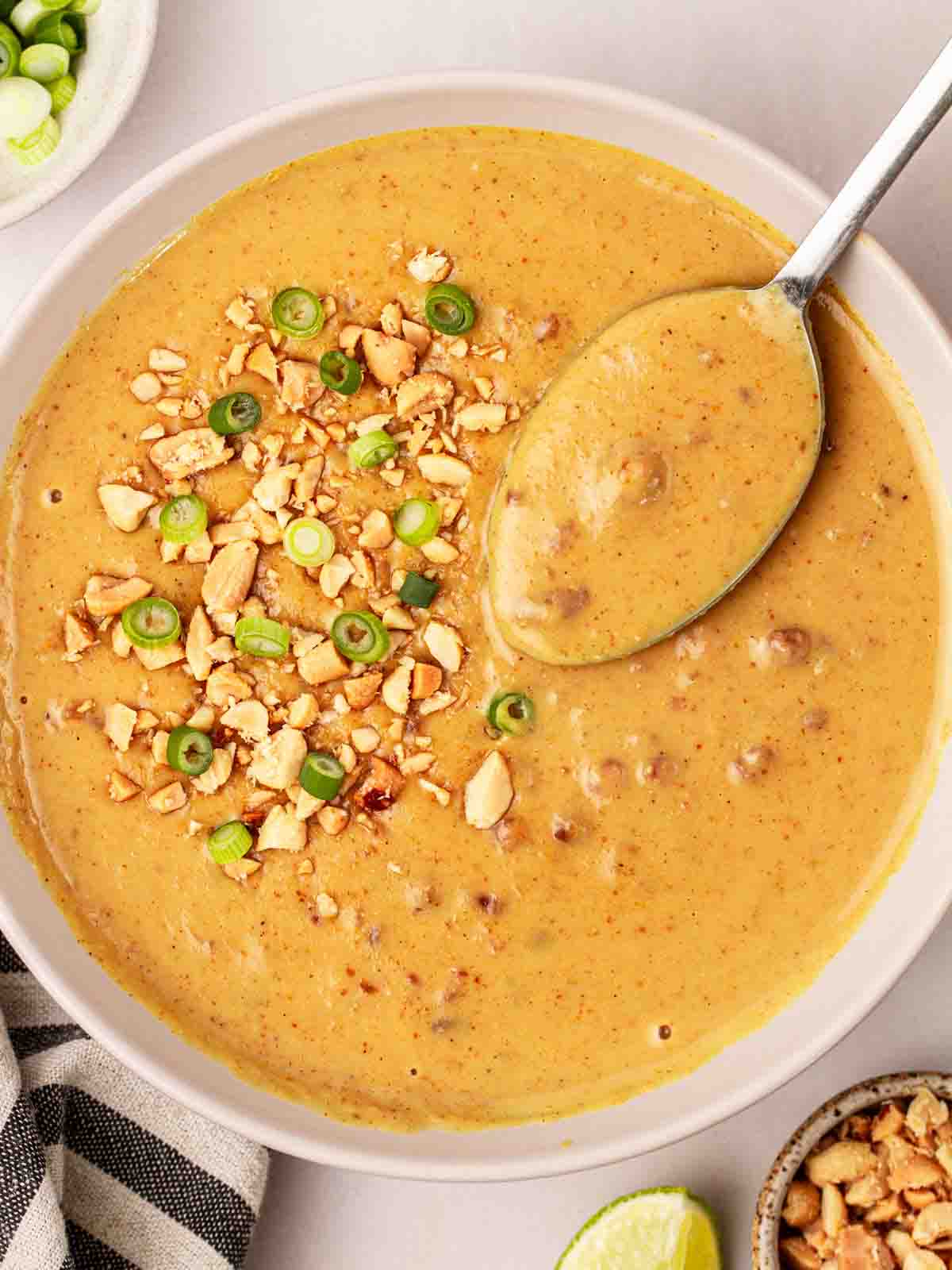 A bowl filled with homemade Satay Sauce, with a spoon to serve. Topped with spring onion and chopped peanuts.